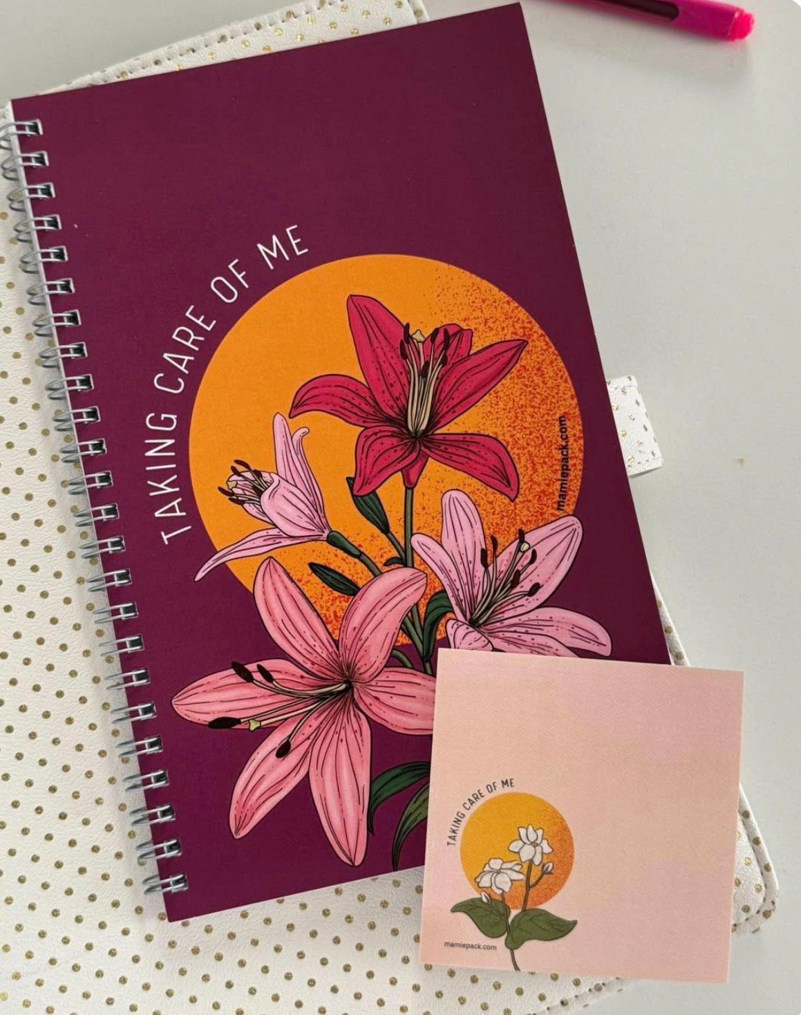 A spiral-bound notebook with a deep purple cover featuring pink lilies and an orange sun graphic with the text 'TAKING CARE OF ME', placed on a white polka-dot surface next to a small matching card and a pink pen.